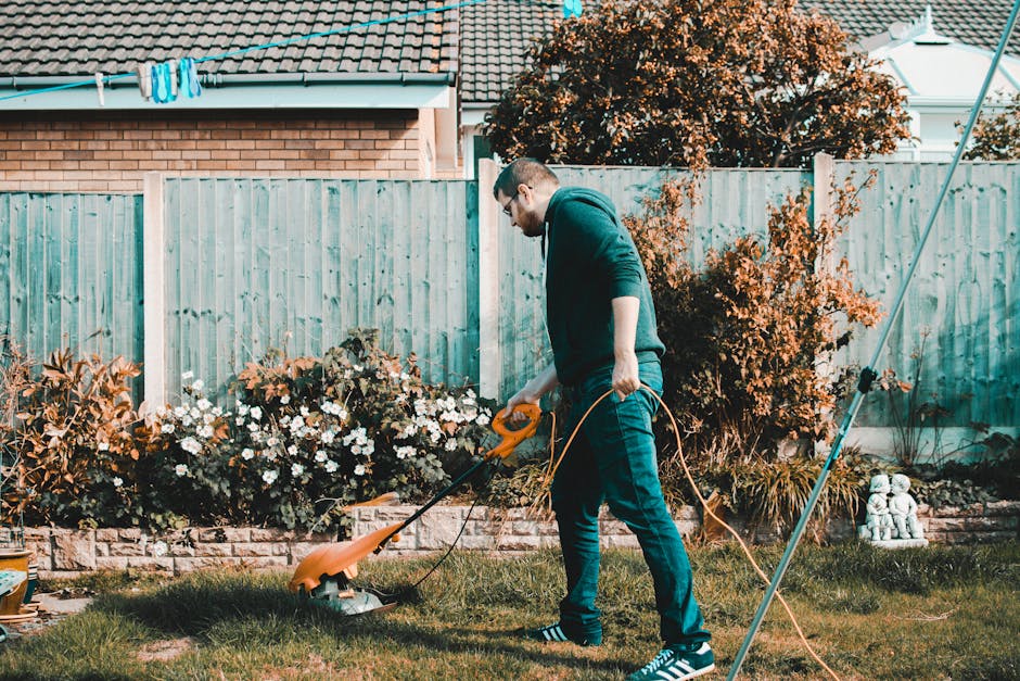 A man with short hair, wearing glasses, a dark hoodie, and blue jeans, is operating a grass trimmer in a well-maintained garden. The garden features a lush green lawn, bordered by flower beds filled with blooming white and purple flowers, and edged with small brick walls. Behind him, there is a wooden fence painted in light blue, with some garden tools and a trellis visible. To the right, a garden rake leans against the fence, and a potted plant rests on the ground. The garden also includes a small dense shrub with reddish-brown leaves, a taller leafy bush, and a large tree with orange foliage in the background, indicating an autumn setting. The scene is lit by soft natural daylight, highlighting the textures of the grass, soil, and plants as part of routine garden maintenance by Gardeners Tooting.