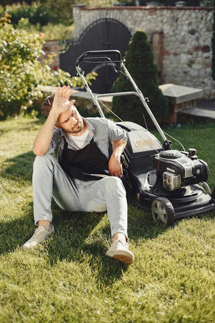 A young man with reddish hair and a beard, dressed in casual gardening clothes including a checkered shirt and light trousers, sits on a well-maintained grassy lawn beside a modern petrol-powered lawn mower. He appears to be resting after working, with his right hand raised to his forehead as if wiping sweat or shielding his eyes from the sun. The garden features lush green grass, neatly trimmed borders, and a stone wall with a black wrought iron gate in the background. There are plants with white flowers on one side, and a small, curated garden bed bordered by soil and stones. Mature trees provide dappled shade, and a stone bench is visible at the edge of the garden. The scene suggests active garden maintenance and landscaping work, supported by the presence of gardening tools and equipment, and is illuminated by natural daylight. The image reflects the outdoor environment managed by Gardeners Tooting, emphasizing garden care and lawn upkeep.