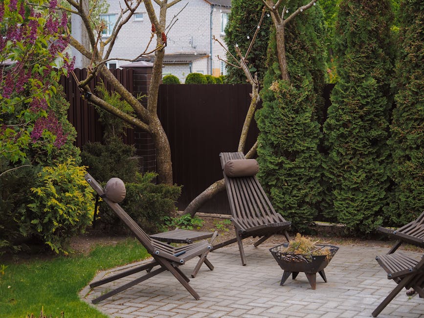 A small backyard garden with a paved patio area featuring three wooden lounge chairs, one of which has a circular cushion attached to the headrest. The garden is enclosed by a dark wooden fence and lush green hedges, including a tall, dense conifer on the right and a flowering shrub with purple blossoms on the left. The space includes a variety of plants, some with textured, glossy leaves and others with softer, lighter foliage. A small, modern firepit with a metallic finish sits on the patio, surrounded by the chairs, creating an inviting outdoor seating area. In the background, a few trees with bare branches extend over the seating area, and neighboring houses with light-colored brick walls are visible behind the fence. Soft, natural lighting suggests a calm, overcast day, highlighting the textures of the garden's plants, soil, and furniture, reflecting typical garden maintenance and landscaping work by Gardeners Tooting.