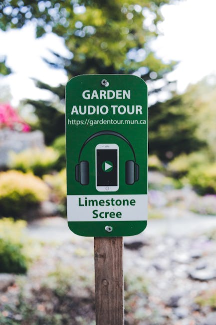 A green garden signage board indicating a garden audio tour for a limestone scree area, mounted on a wooden post within a well-maintained front garden. The garden features a neatly trimmed lawn with lush, green grass, bordered by garden beds planted with colorful flowers, shrubs, and low hedges. In the background, mature trees provide partial shade, and a paved pathway leads through the space. The environment appears sunny with bright lighting, highlighting the textures of the soil, foliage, and gravel. This setting showcases professional garden maintenance and landscaping work, with tools and trimmed plants visible, emphasizing the care taken by Gardeners Tooting in creating an inviting outdoor space suited for local gardeners exploring front garden planting.
